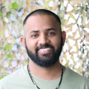 Stock photo showing headshot of Indian man with shaved head wearing t-shirt and necklaces.